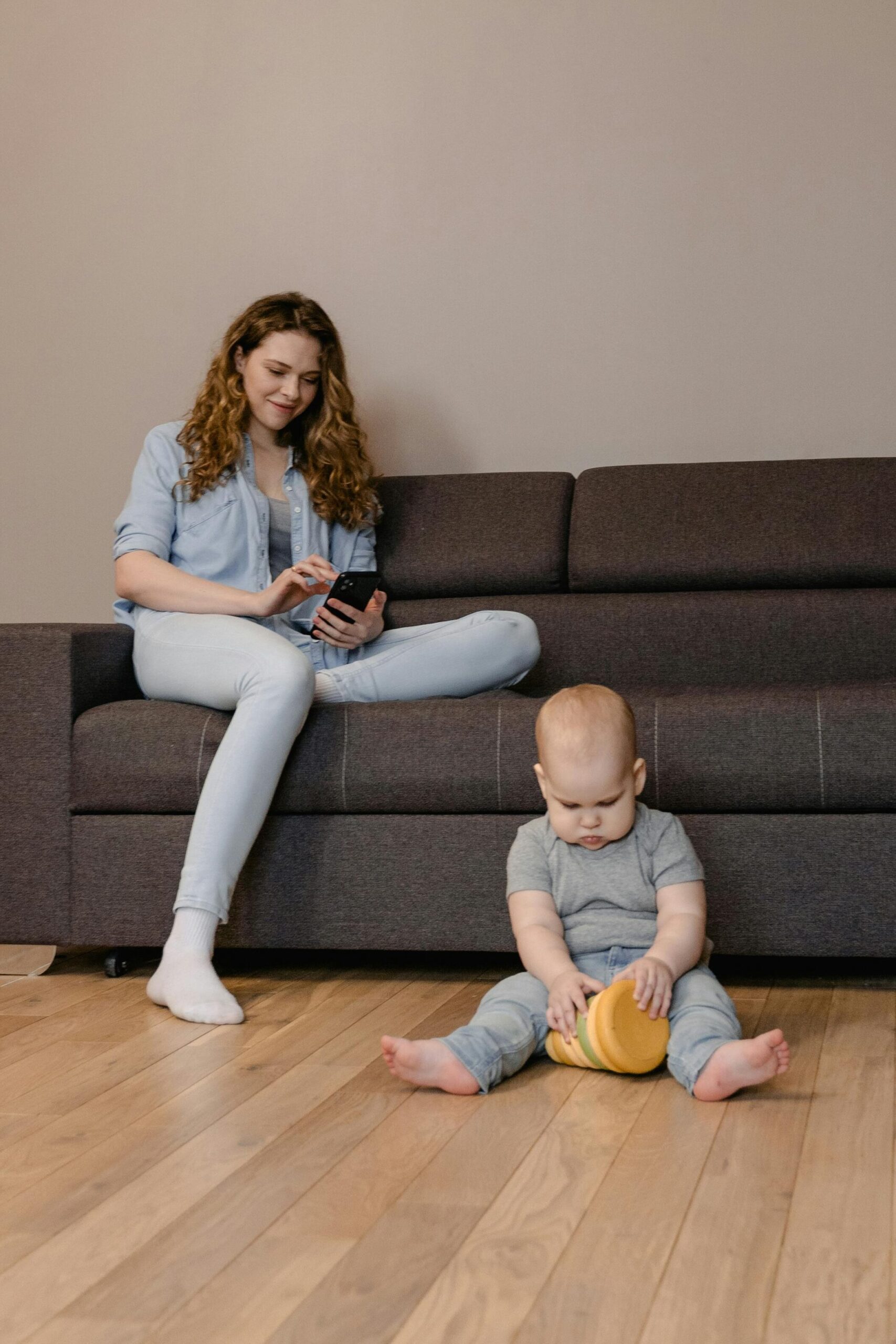 A Woman Sitting on the Couch Using Her Phone while Her Baby is Sitting on the Floor